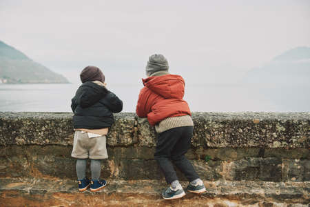 Outdoor portrait of two funny kids playing by the lake on a cold autumn day, wearing warm winter jackets, back viewの写真素材