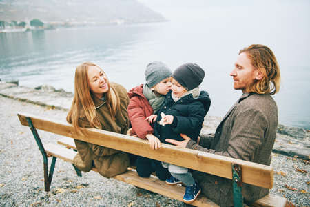 Family of 4 sitting on bench, enjoying nice autumn day by the lake, cold weatherの写真素材