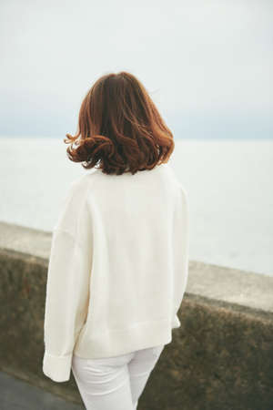 Outdoor portraiit of beautiful woman wearing white pullover, relaxing next to lake or sea, back viewの写真素材