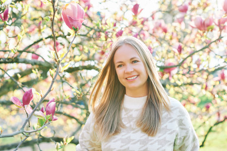 Outdoor portrait of beautiful happy model with blond hair posing next to magnolia flowersの写真素材