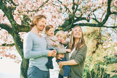 Outdoor portrait of happy young family playing in spring park under blooming magnolia tree, lovely couple with two little children having fun in sunny gardenの写真素材