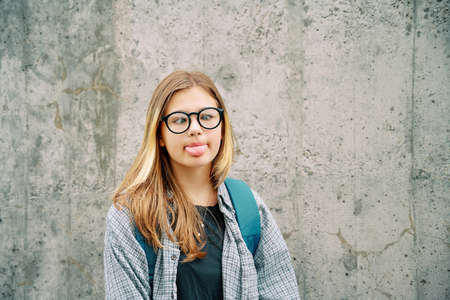 Outdoor portrait of silly young teenage kid girl pulling a tongue, crossing eyes over her nose, wearing glasses and backpack, posing on grey wall backgroundの写真素材