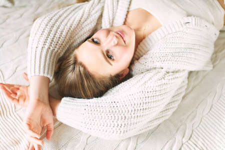 Portrait of young girl lying on white blanket, looking straight at camera, wearing warm knitted cardiganの写真素材