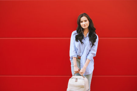 Outdoor portrait of young pretty woman posing on red background, holding backpackの写真素材