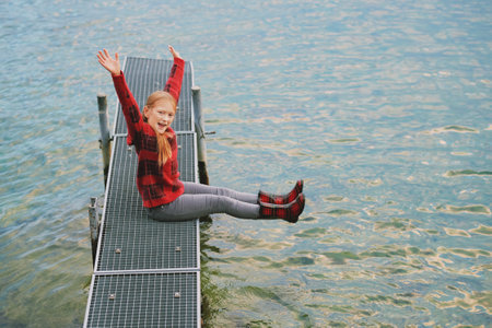 Outdoor portrait of cute little girl posing next to lake on dock, wearing red sweater and rain...