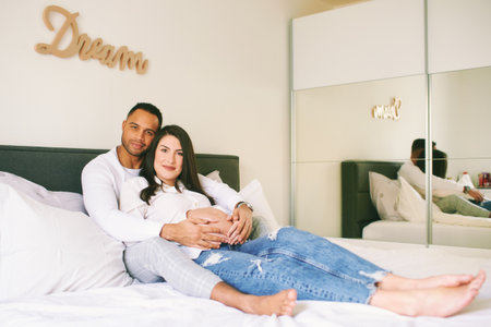 Indoor portrait of happy young family, pregnant woman with her loving husband relaxing on bedの写真素材