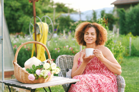 Outdoor portrait of beautiful mature woman resting in summer garden, sitting in cozy chair, holding cup of tea or coffee, basket with freshly cut rosses standing on the tableの写真素材