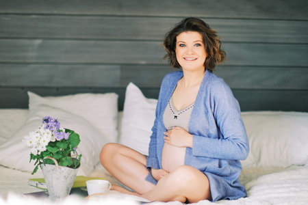 Early morning portrait of happy young pregnant woman resting in bed, drinking tea