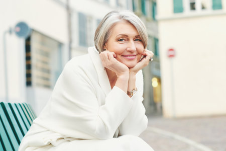 Outdoor portrait of beautiful and elegant middle age 55 - 60 year old woman, wearing white jacket, sitting on bench outsideの写真素材
