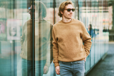 Outdoor portrait of handsome young man wearing sunglasses and beige fuzzy fleece sweater, posing next to mirror wall on city street backgroundの写真素材