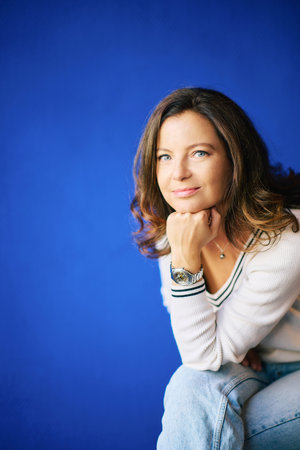 Portrait of pretty 40 year old woman posing next to bright blue wall, sitting and leaning on hand