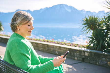 Portrait of middle age woman enjoying nice day outside, relaxing on bench in front of mountain lake, holding phone, reading or texting messageの写真素材