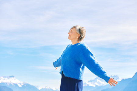 Outdoor portrait of happy middle age 55 - 60 year old woman with arms open wide, posing on blue sky and mountain backgroundの写真素材