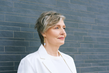 Headshot portrait of 55 - 60 year old woman with gray hair, wearing white jacket, posing on gray backgroundの写真素材