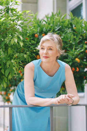 Outdoor summer portrait of charming 50 - 55 year old woman wearing blue dress, posing outside next to potted plant treeの写真素材