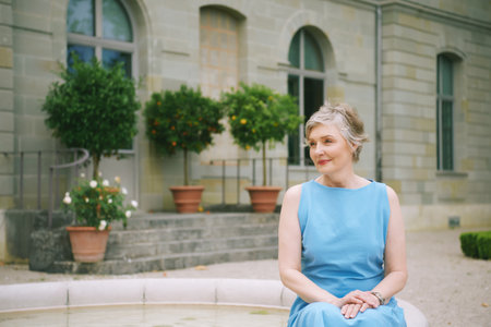 Outdoor portrait of mature woman of 50 - 55 years old, wearing blue dress, relaxing next to fountain...