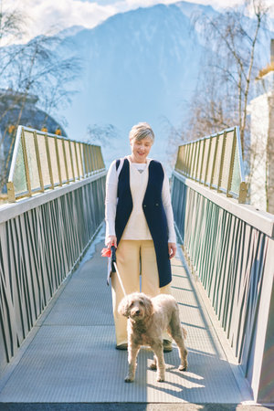Outdoor portrait of happy mature woman walking with labrodoodlee dog, posing on bridgeの写真素材