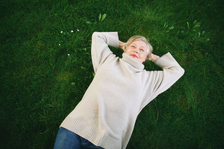 Outdoor portrait of happy mature woman laying on green grass, wearing warm pullover or sweater....
