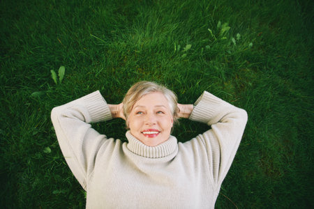 Outdoor portrait of happy mature woman laying on green grass, wearing warm pullover or sweater....