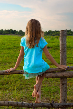 A little girl without shoes sits on a wooden fence in a meadow in summerの写真素材