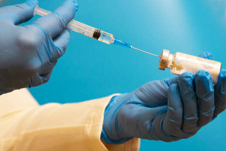 Doctor filling syringe with medication, closeup. Vaccination and immunization. a doctor wearing gloves with an ampoule and a syringe on a blue backgroundの写真素材