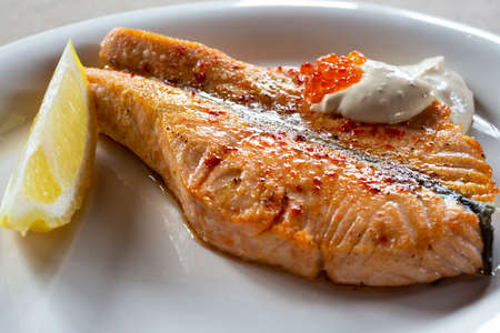 A glass of red wine and grilled salmon with white sauce, red caviar and lemon on a white plate in the restaurantの写真素材