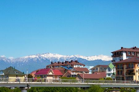 Pedestrian bridge over the Mzymta river in the resort town, view of colorful houses and snow-capped mountains against the blue sky. Adler, Sochi, Russia - May 15, 2022のeditorial素材