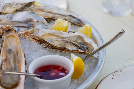 Fresh oysters on the halves of the shell are served in a plate with ice, lemons and sauce.