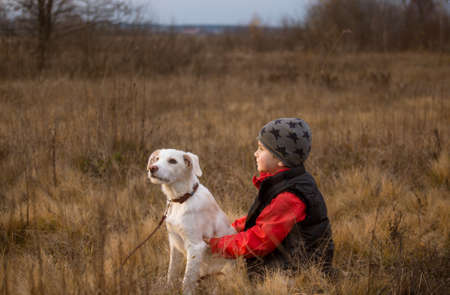todler boy walks in nature with his white young retriever dog, sit in an embrace among dry grass. The joy of communicating with a four-legged friend, a beloved petの写真素材