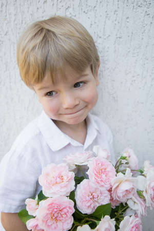 Portrait of a smiling cute toddler boy with a bouquet of pink rose flowers. Congratulations for mom. Festive bouquet with love. Gift for mother's day, birthday. Tenderness, harmony, calmness, joyの写真素材