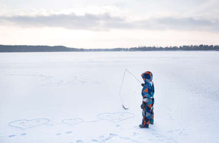 boy in a multi-colored winter overalls in an open snow-covered space, with a stick in the snow, draws hearts, enjoys the winter. Winter fun in nature. Love winterの写真素材