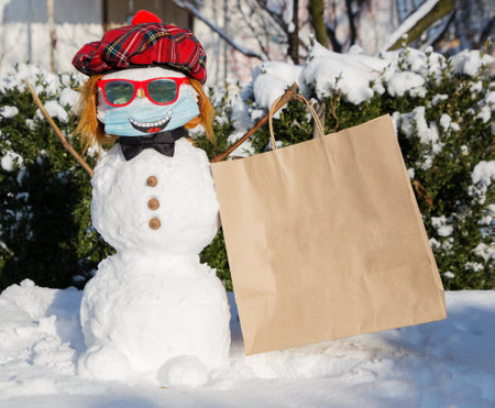 happy snowman in a bright red beret, sunglasses and a medical mask with a smile painted on it holds a large craft bag for the inscription of advertising text on it.の写真素材