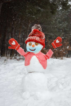 happy snowman in red knitted hat, sweater, mittens, medical mask with smile painted on it stands in snowy forest.の写真素材