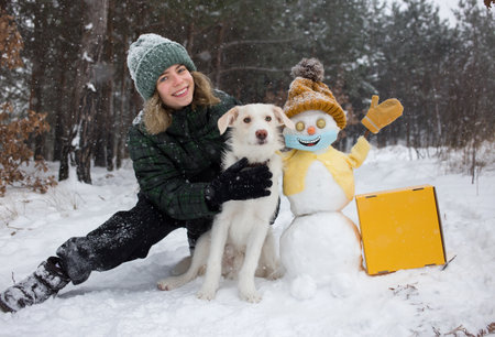 Happy cute teenager girl, white dog, cheerful snowman in yellow knitted hat, jacket, mittens, medical mask with smile painted on it in a snowy forest. life during pandemic. Winter activitiesの写真素材