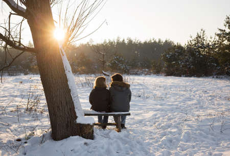 young woman and a man are sitting on a bench with their backs in the frame in a secluded place, enjoying each other and winter sunset. good relationship, falling in love, romance, tender feelingsの写真素材