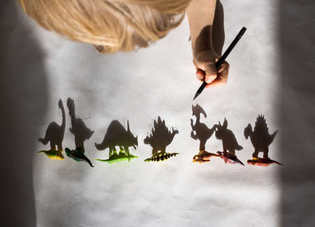 preschooler draws with pencil contrasting shadows from toy little dinosaurs standing in a row. drawing of child, creative ideas for children's creativity. light and shadow. Selective focus.の写真素材