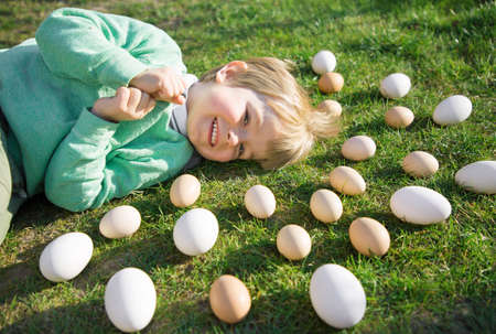 many white and beige chicken and goose eggs lie on the bright green grass around the head of a cheerful, happy 3-4 year old blond boy. On the farm, preparing for Easter. Cheerful atmosphereの写真素材