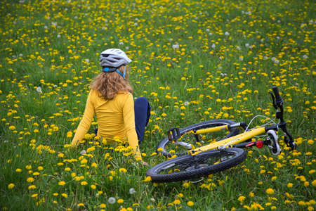 unrecognizable teenage girl in a bicycle helmet sits with her back on green grass among yellow dandelions, bicycle lies nearby. cycling in nature, harmony, energy of nature, active healthy lifestyleの写真素材