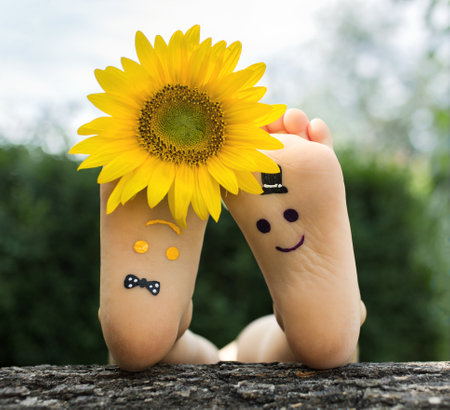 Yellow and purple upside down funny faces are painted on the bare feet of a child lying on the grass. Sunflower flower between fingers, cheerful positive atmosphere, happy childhood, life with joyの写真素材