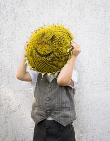 unrecognizable child holds in front of face large yellow sunflower head, on which muzzle with smile...