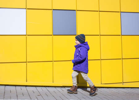 Toddler boy in a purple jacket walks forward along the walls of a bright yellow wall. step forward...