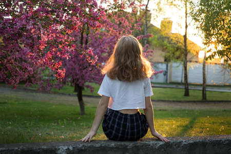 unrecognizable young woman sits with her back near a large blooming pink spring tree. Dreams, enjoys the moment. Digital detox. harmony of human and natureの写真素材