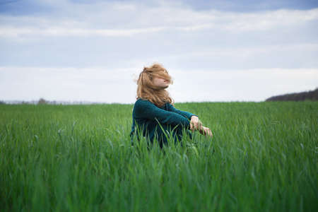teenage girl in sweater and long emerald-colored skirt sits on ground among field of young grass, her face is covered with hair that flutters in wind. Harmony, inspiration, relaxation, self-isolationの写真素材