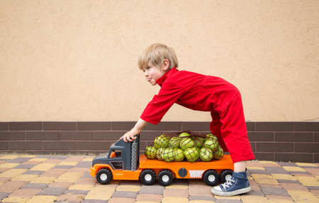 positive toddler boy in a red overalls carries a lot of organic farm apples on a big toy car - a truck. Courier, truck driver, like dad. games for boysの写真素材