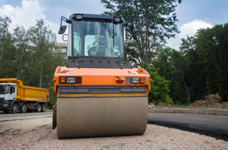 large orange roller on road paving works. In the background is a dump truck. Improvement of the...