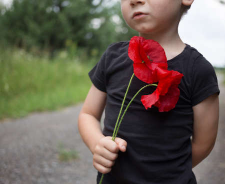 sad toddler boy in a black T-shirt holds two poppy flowers in his hands. Focus on the flower. Reflection on the topic of war. Memorial dayの写真素材