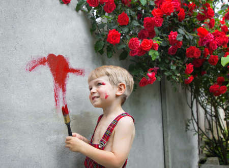 cute toddler boy in overalls draws a heart with red paint on a concrete fence near a blooming bush of red roses. Valentine's Day, surprise gift for mom. Summer funの写真素材