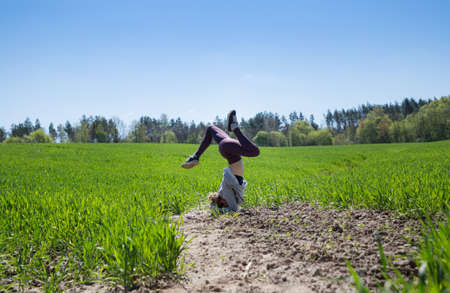 young slender woman stands on her head upside down, holding her balance, among the bright green...