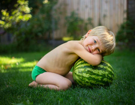 On a hot summer day, a cute barefoot toddler boy without clothes sits on the grass next to a large striped watermelon. Waiting for an appetizing delicious dessert. Cheerful carefree childhoodの写真素材