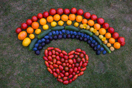 bright ripe multicolored vegetables and fruits - tomatoes, cucumbers and plums are laid out on a green lawn with a multicolored rainbow, a heart of red tomatoes is laid out under the rainbowの写真素材